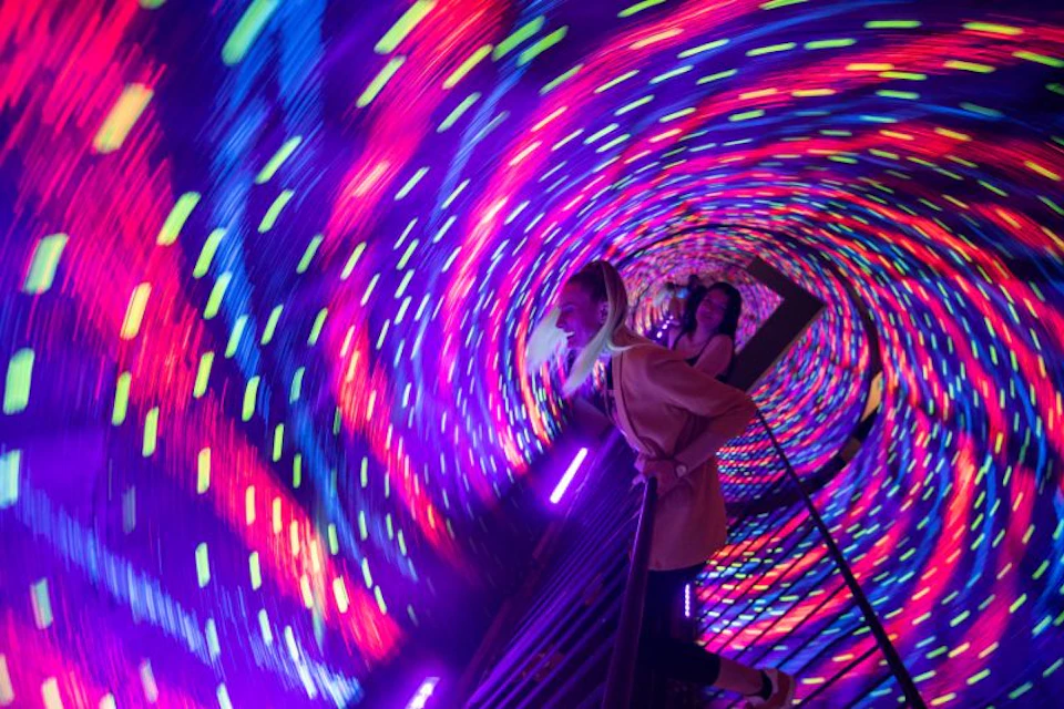 Two people walk through a tunnel with vibrant, swirling neon lights. One leans on the railing, looking forward.