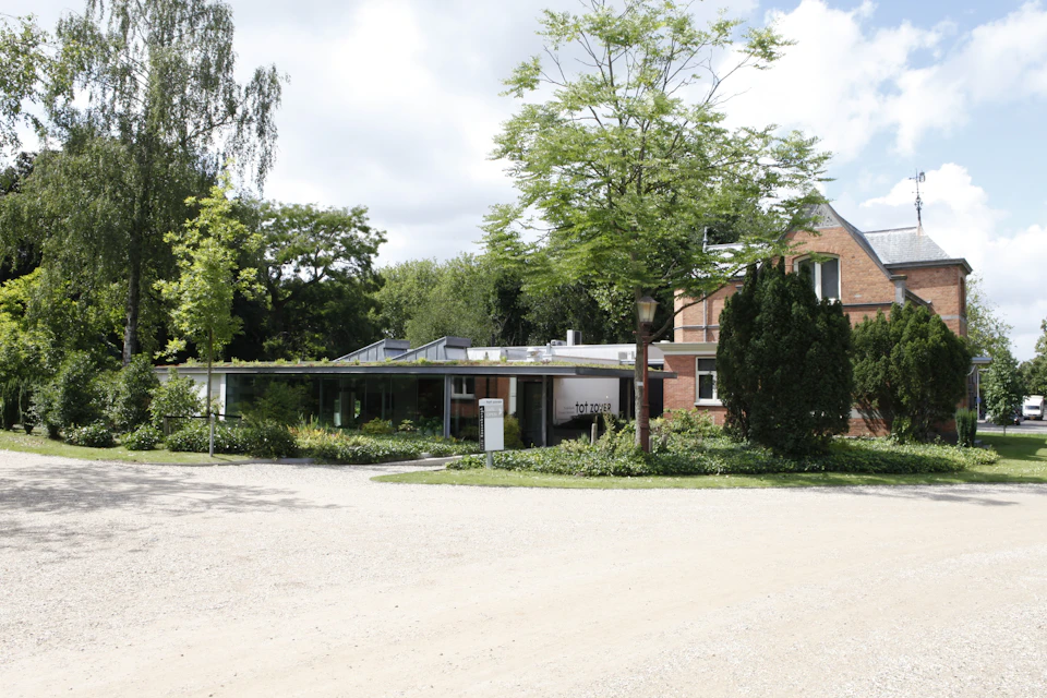 Modern building with glass walls and a flat roof, surrounded by trees and greenery, next to a traditional brick house.