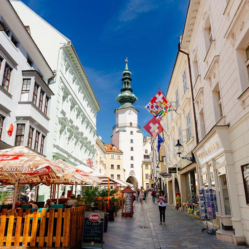En livlig smal gata med uteserveringar, färgglada byggnader, ett klocktorn och olika flaggor, under en klarblå himmel.