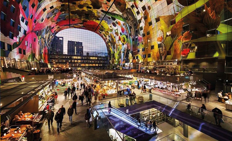 A bustling indoor market with colourful art on the arched ceiling, food stalls, shoppers, and an escalator.