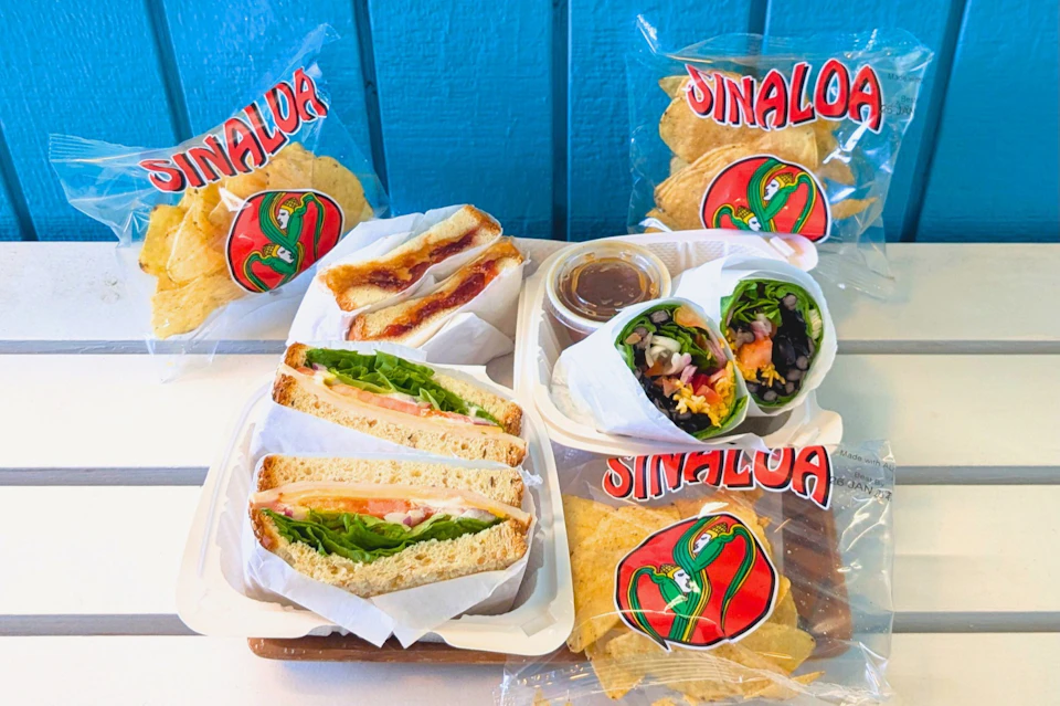 Assorted sandwiches, wraps, and chips in branded "Sinaloa" bags on a table. Blue background.
