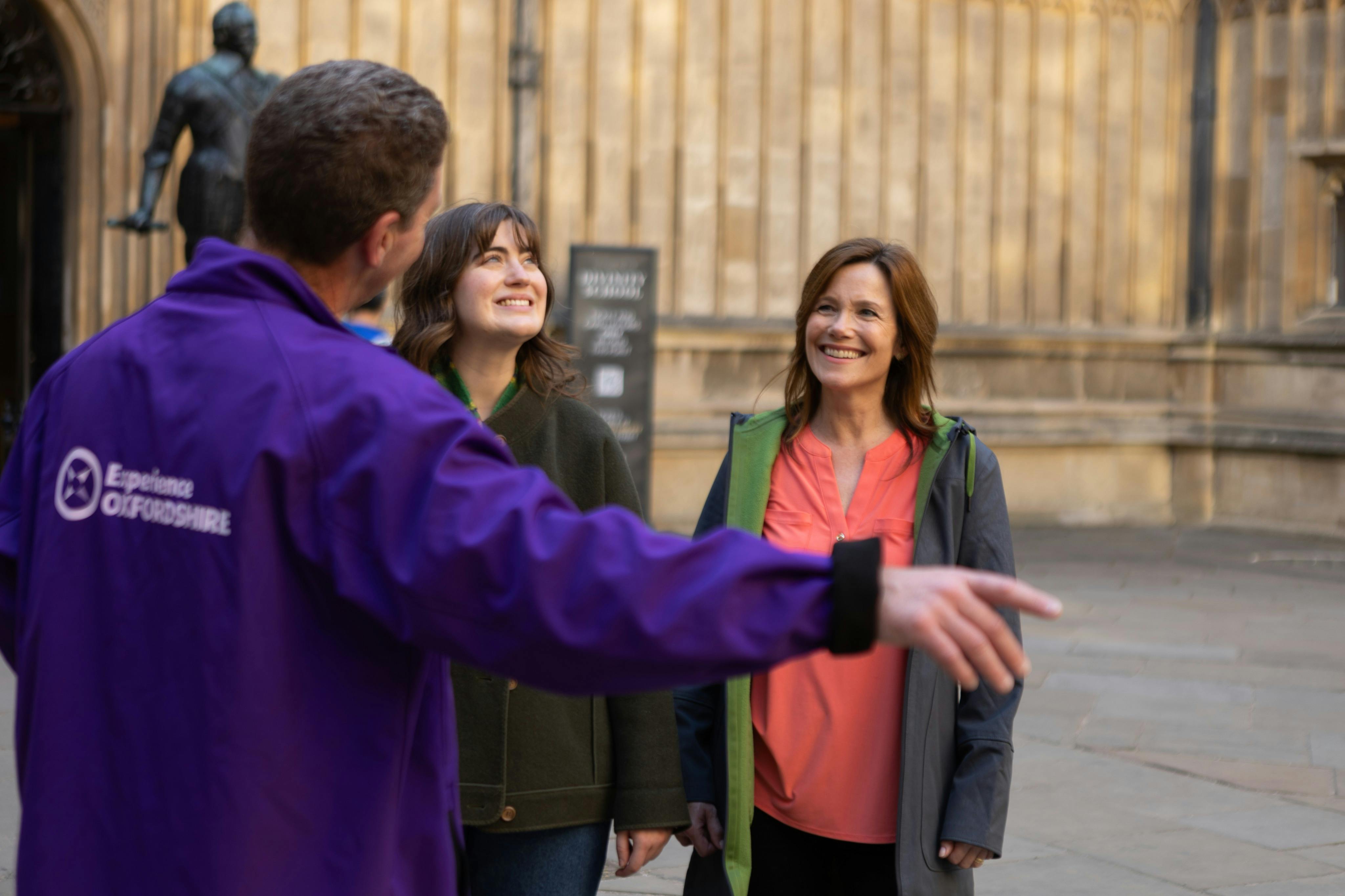 A man in a purple jacket gestures while speaking to two women smiling outdoors near a beige wall.