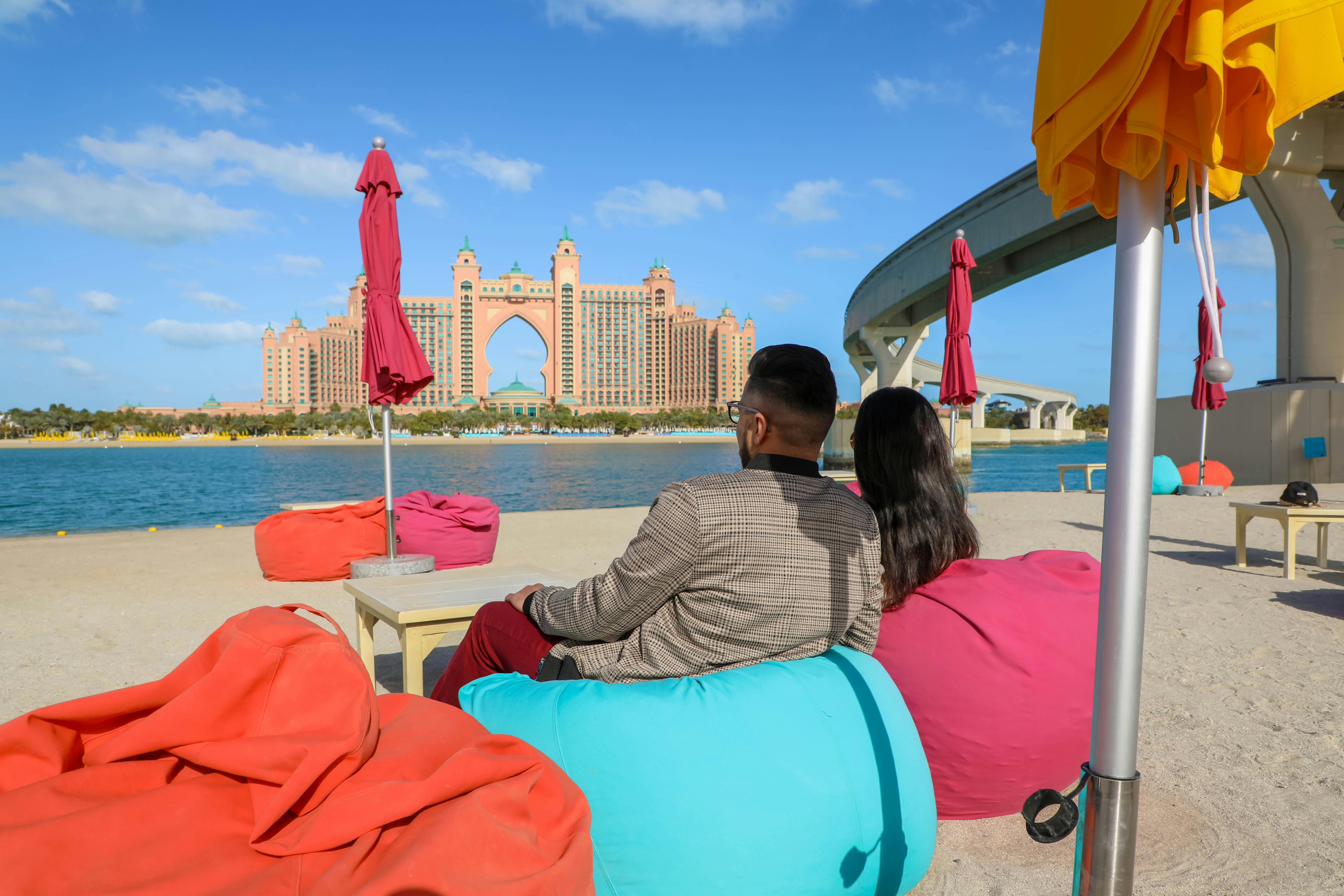 Due persone siedono su sacchi di fagioli colorati vicino all'acqua, di fronte a un grande hotel rosa con archi, sotto un cielo blu con nuvole sparse.