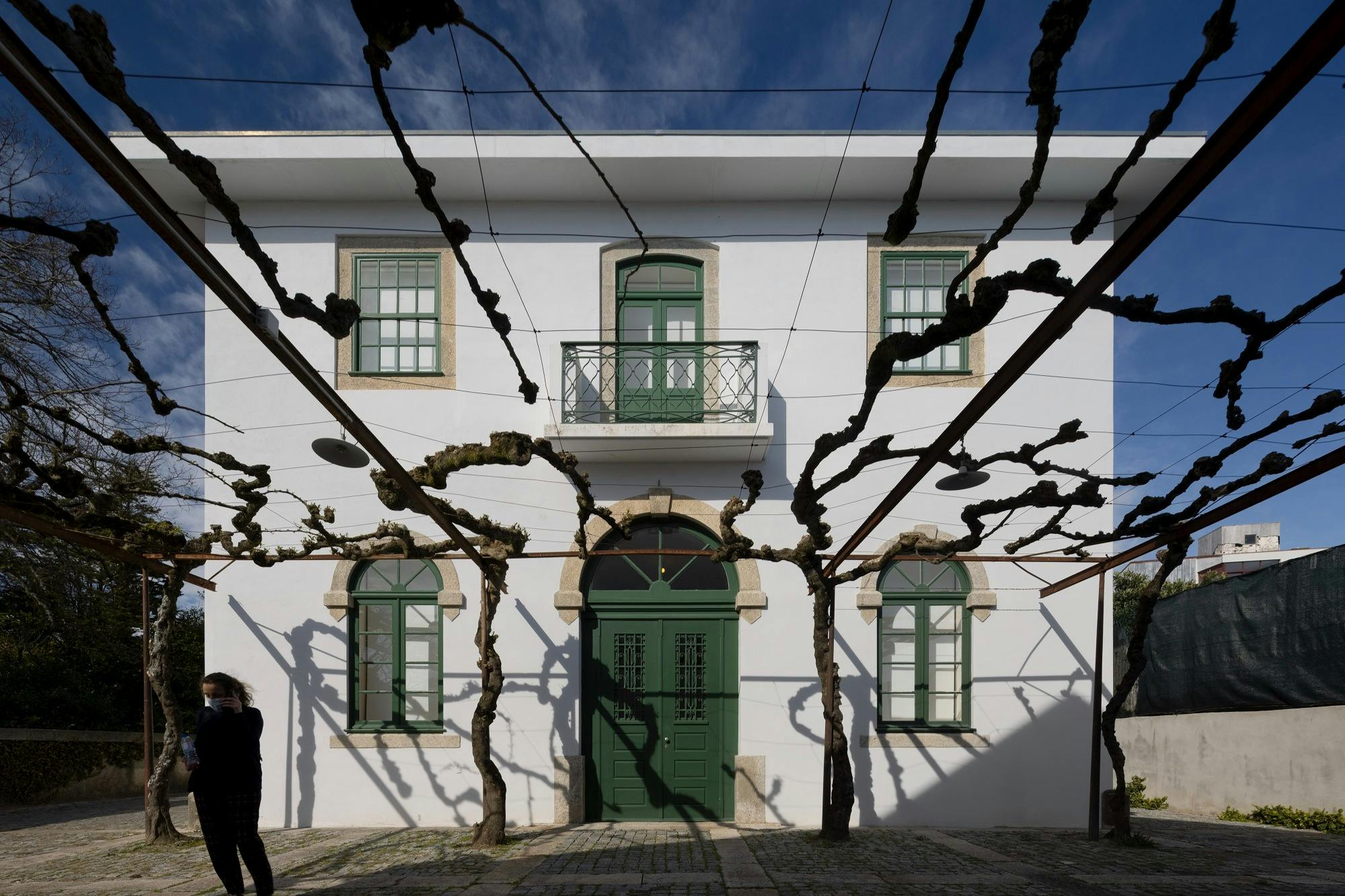 A white building with green doors and windows, bare tree branches, and a cobblestone pathway. One person is partially visible.