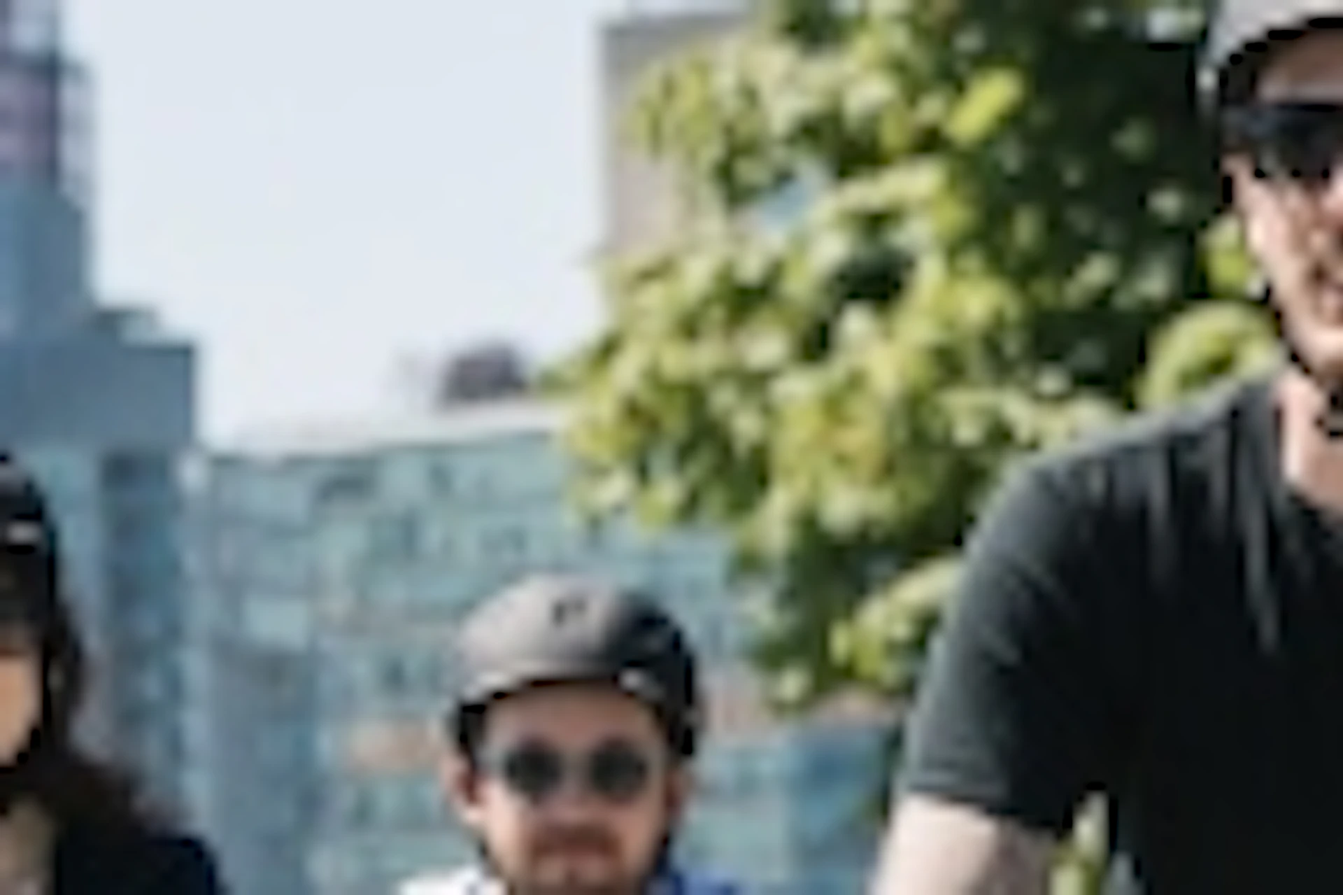 Four people biking on a path in a park, with tall city buildings and greenery in the background.