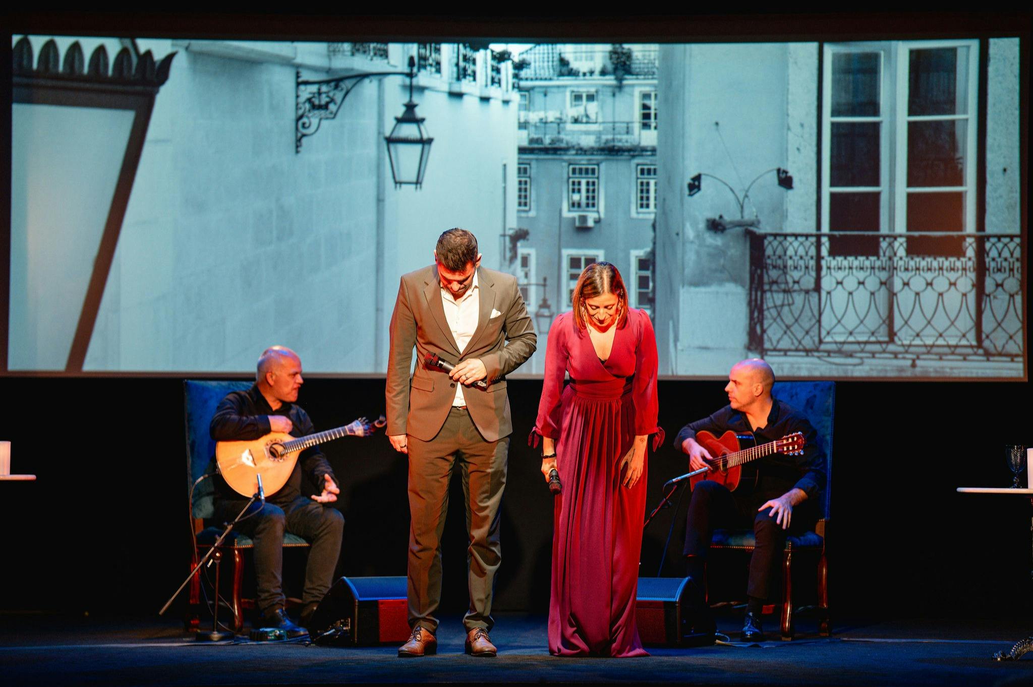 Two performers bowing on stage, dressed formally. A guitarist sits behind them, and a black-and-white cityscape is projected in the background.