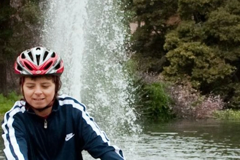 Three people with helmets ride bicycles near a fountain and a neoclassical building with columns and a dome.