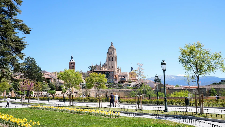 People walking by a landscaped park with trees and flowers, with a historic cathedral and town in the background under a clear blue sky.