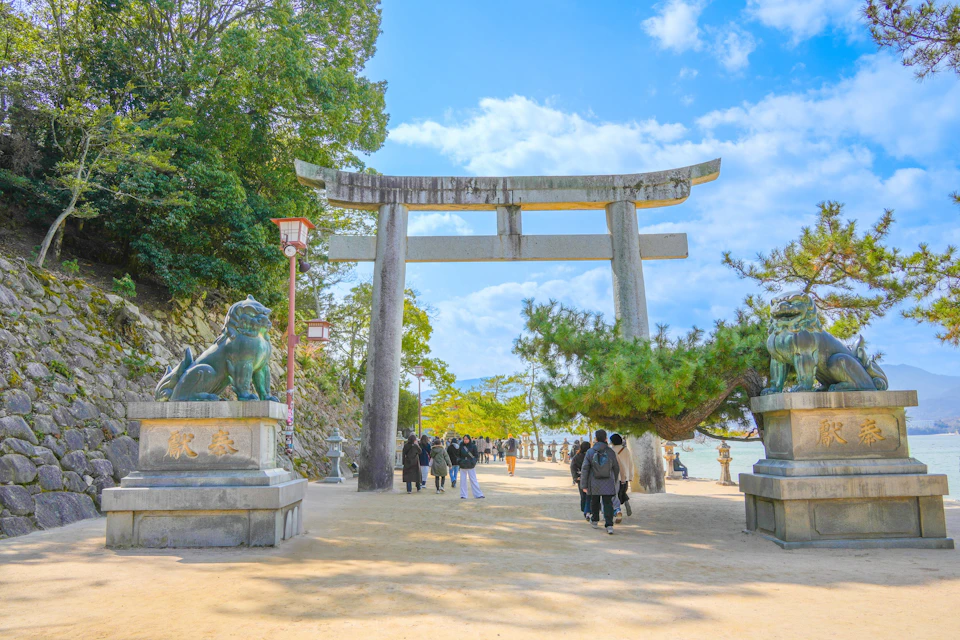 People walking under a large stone torii gate flanked by guardian statues, set in a scenic outdoor area with trees and a bright sky.