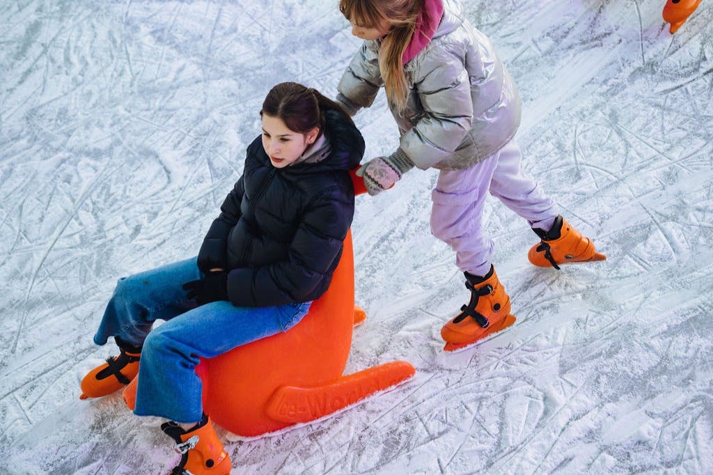 Twee mensen op een ijsbaan. De een zit op een oranje ijsslee en de ander duwt van achteren. Beiden dragen winterkleding en schaatsen.