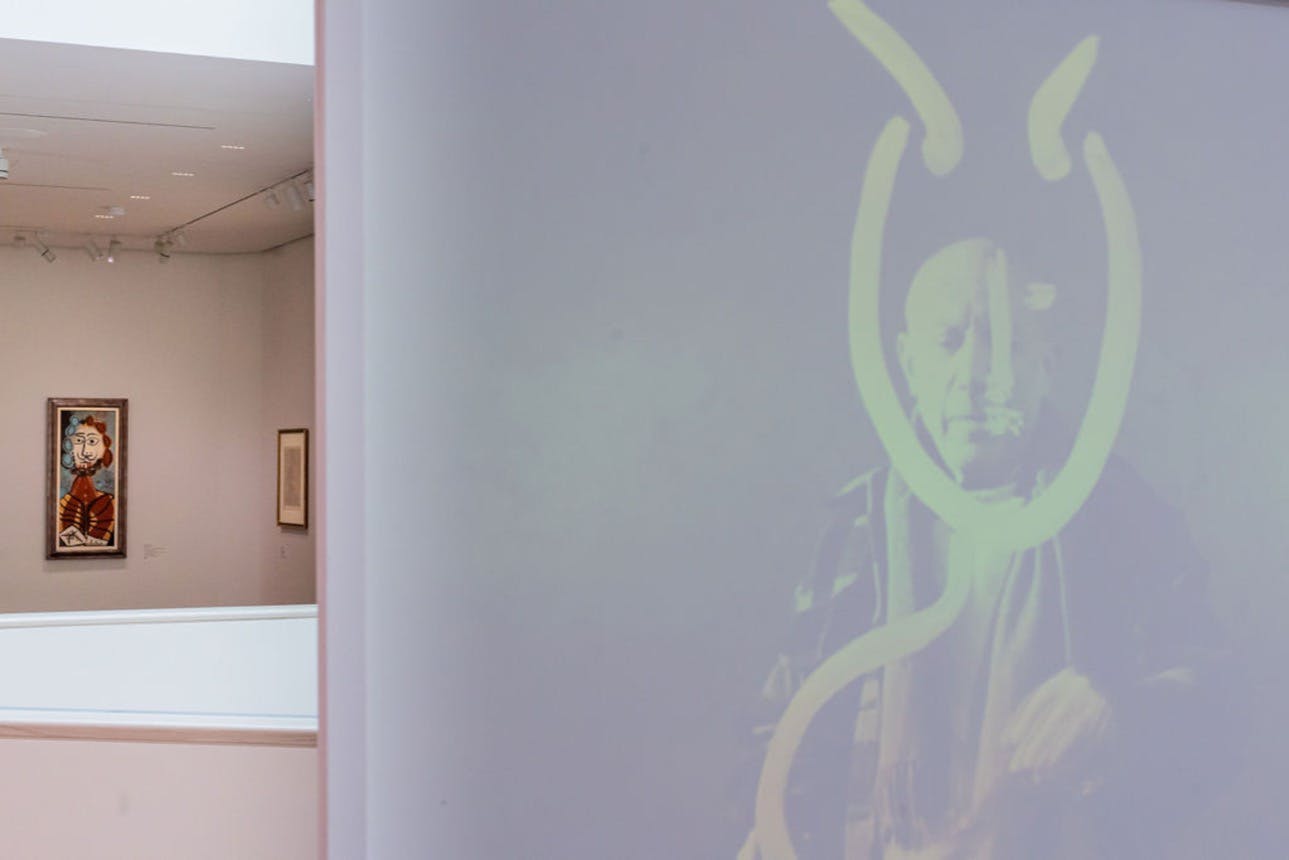 Špatně osvětlená místnost s částečně viditelným promítaným obrazem mužské tváře, orámovaná abstraktními liniemi. Závěsné umělecké dílo v pozadí.