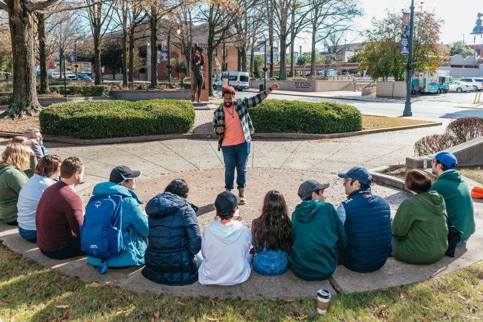 A person speaks to a seated group of people in an outdoor park with trees, bushes, and a statue in the background.