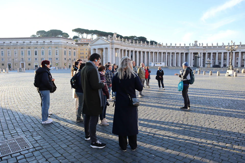 A group of people listening to a guide in an outdoor plaza with columns in the background, under a clear blue sky.
