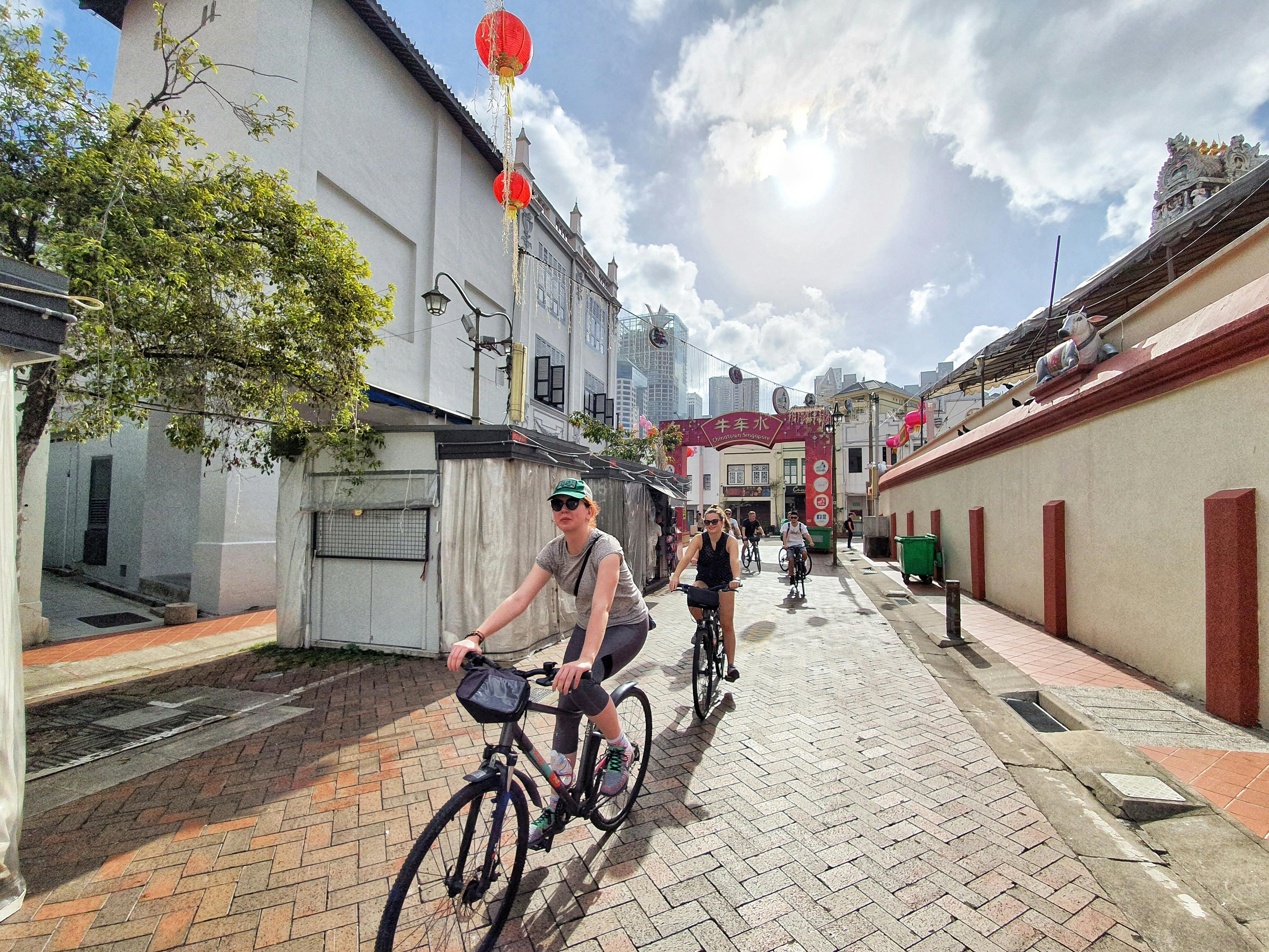 Menschen auf Fahrrädern auf einer engen, sonnenbeschienenen Straße, flankiert von Gebäuden mit roten Laternen und einem Tor im Hintergrund.