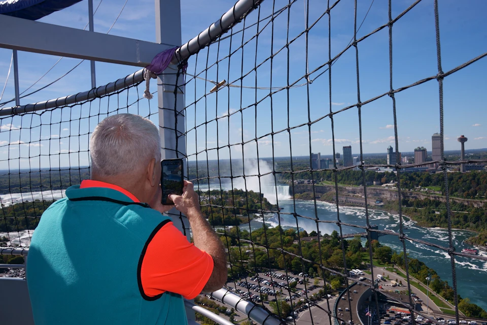 A person photographs Niagara Falls through a safety net from an elevated viewpoint on a sunny day.