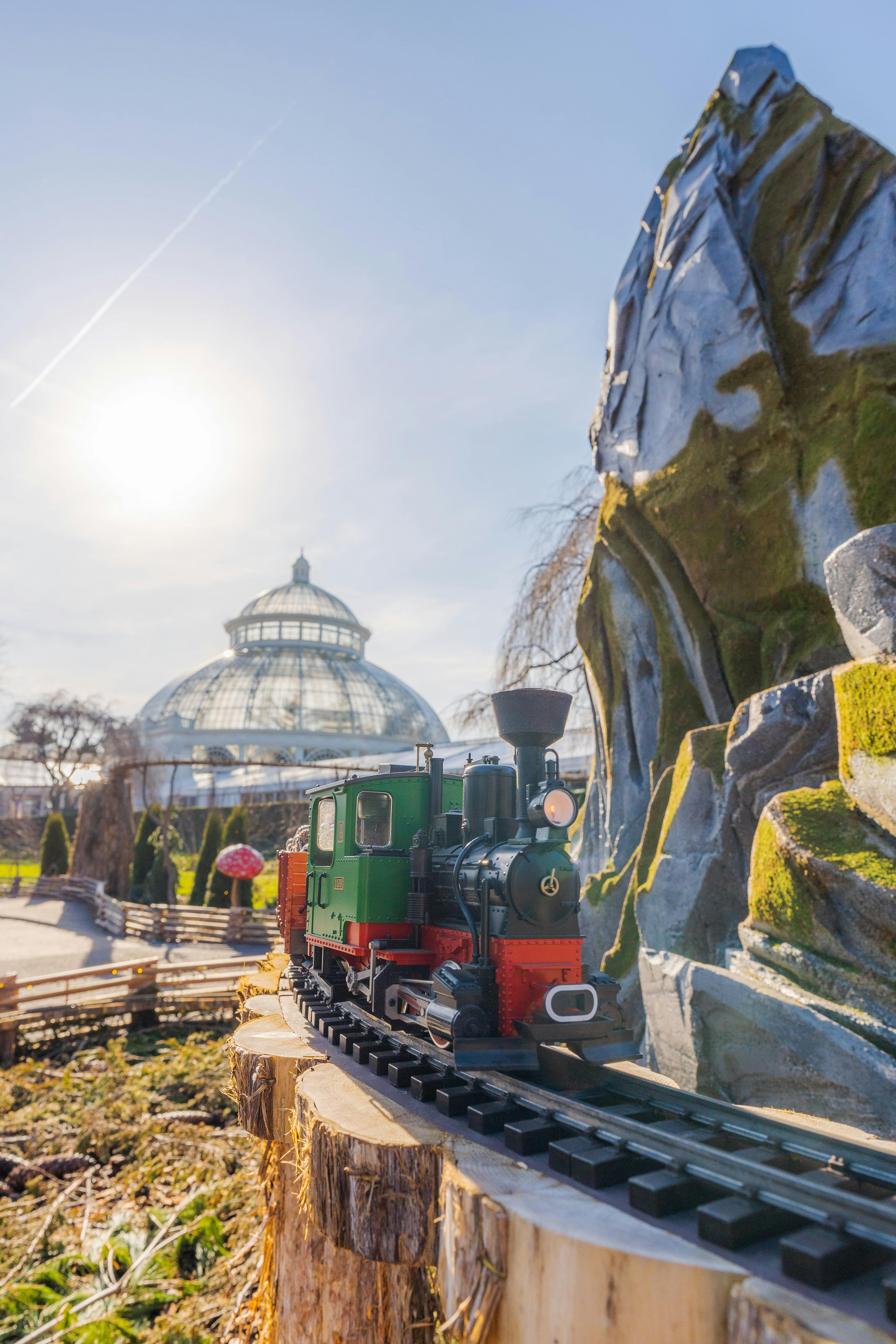 Et legetøjstog på spor ved siden af en stenet struktur med en udestue med glaskuppel og en solrig himmel i baggrunden.