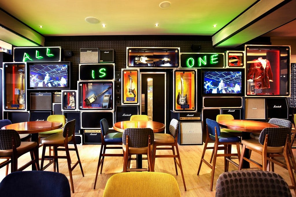 테이블, 의자, 기타, 앰프, 네온사인, TV 화면이 있는 벽면 디스플레이가 있는 세련된 바입니다.