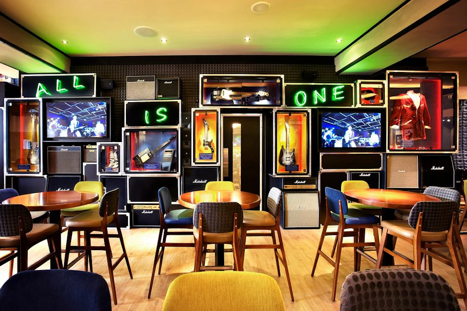 A stylish bar with tables, chairs, and a wall display featuring guitars, amplifiers, neon signs, and TV screens.