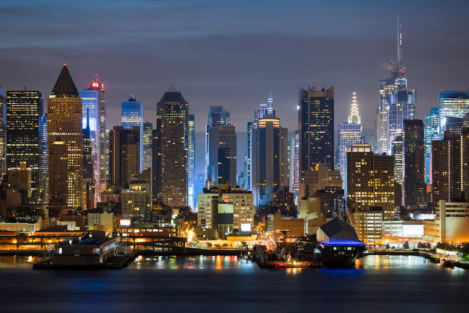 A nighttime cityscape featuring illuminated skyscrapers along a waterfront, with reflections of the buildings in the water.