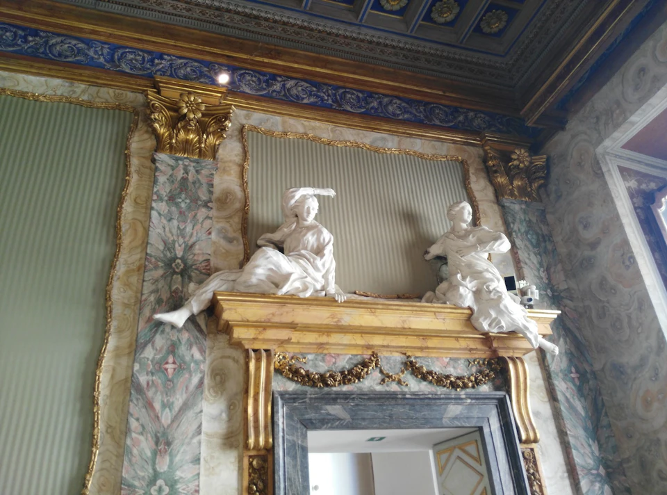 Two white statues of robed figures are mounted above an ornate gold-framed doorway in an elaborately decorated room.