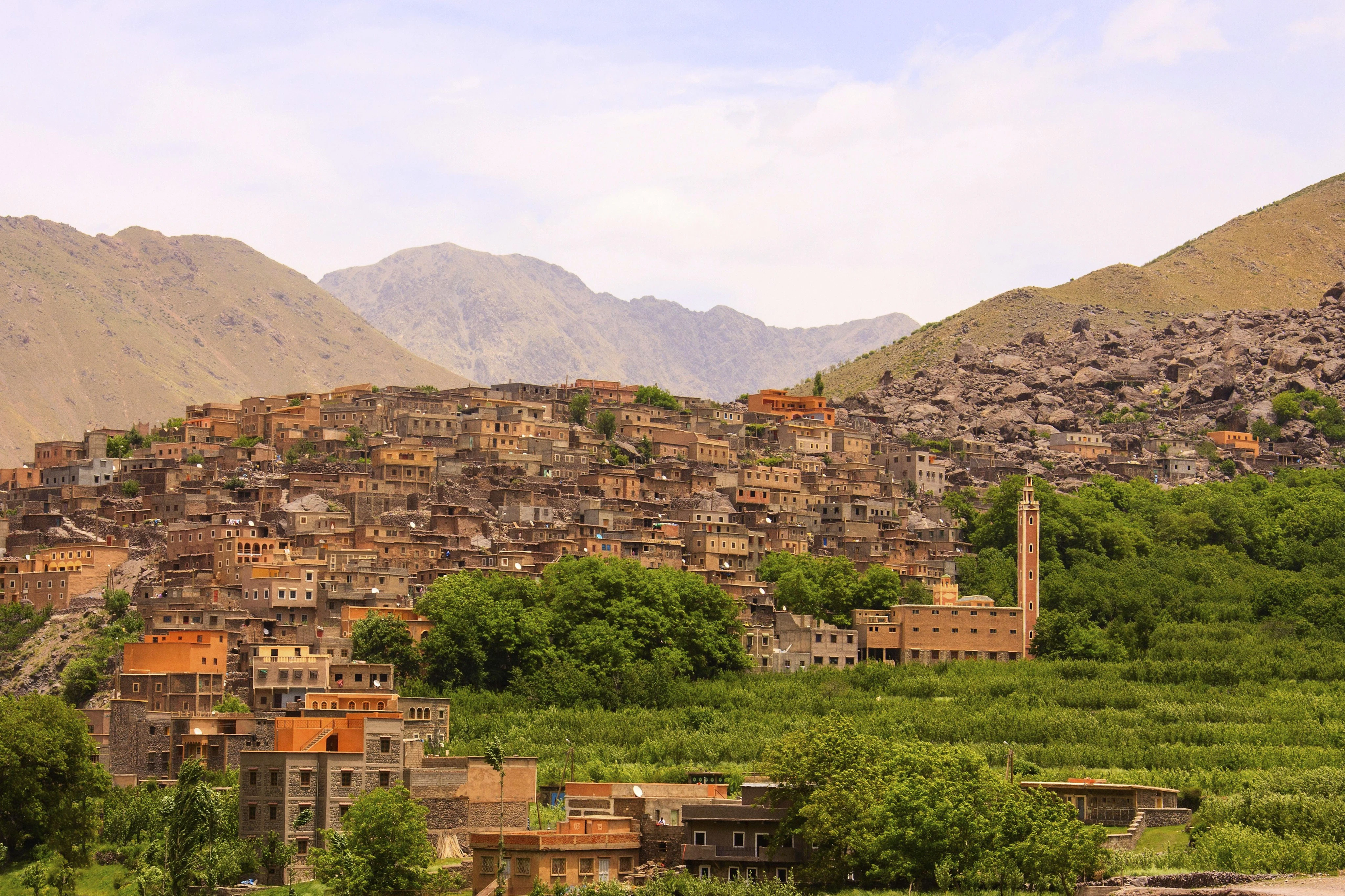 Mountain village with densely packed brick houses, green trees in the foreground, and rocky hills in the background under a cloudy sky.