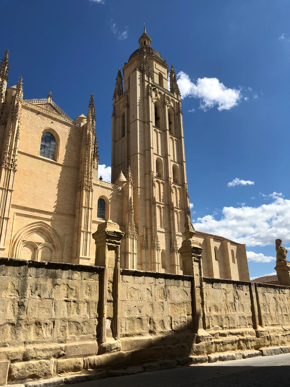 A tall, historic stone cathedral with intricate details, set against a clear blue sky with scattered clouds, viewed from behind a stone wall.