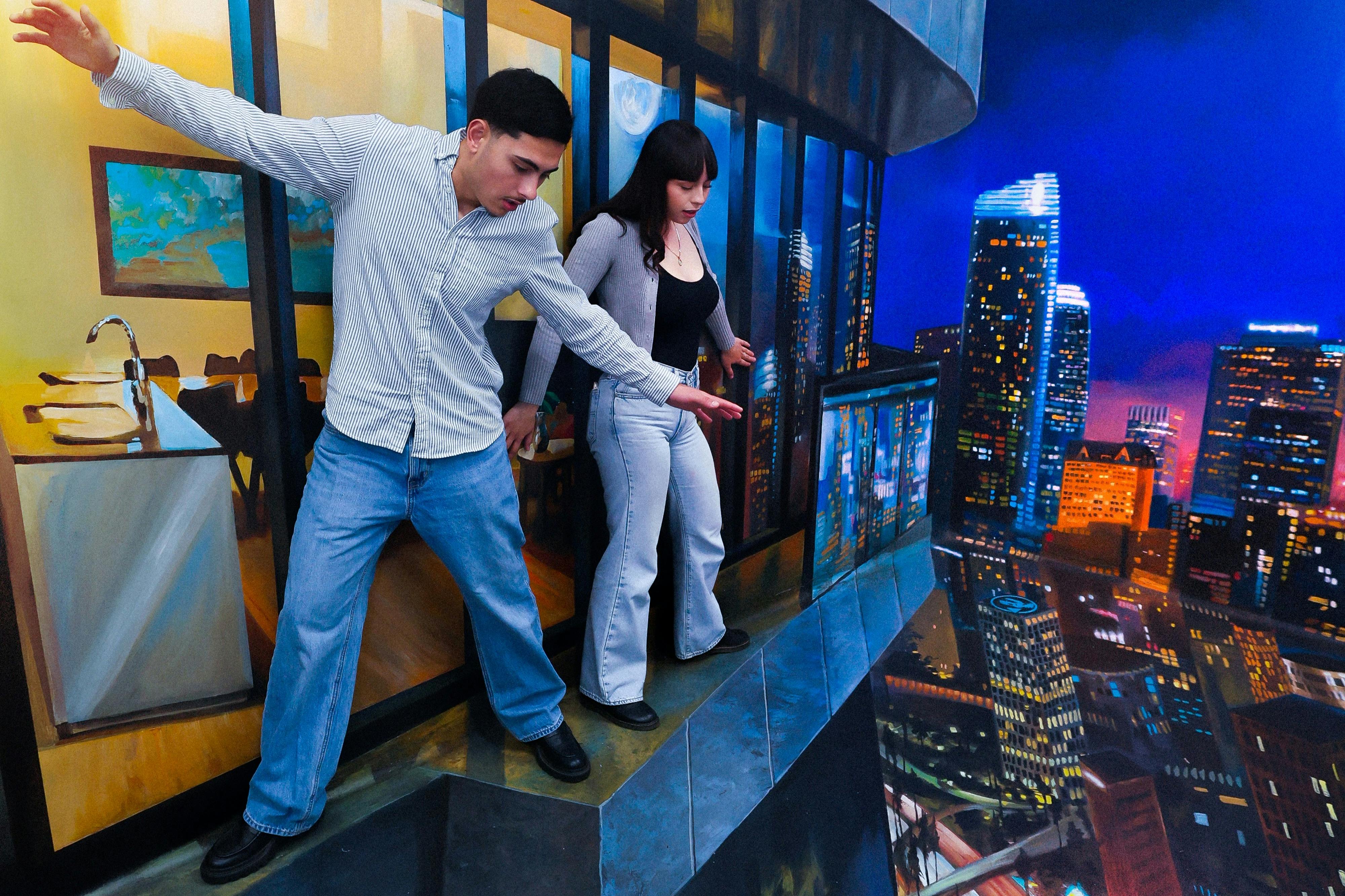 Two people carefully walking on a ledge painted to resemble a cityscape at night with a kitchen interior in the background.