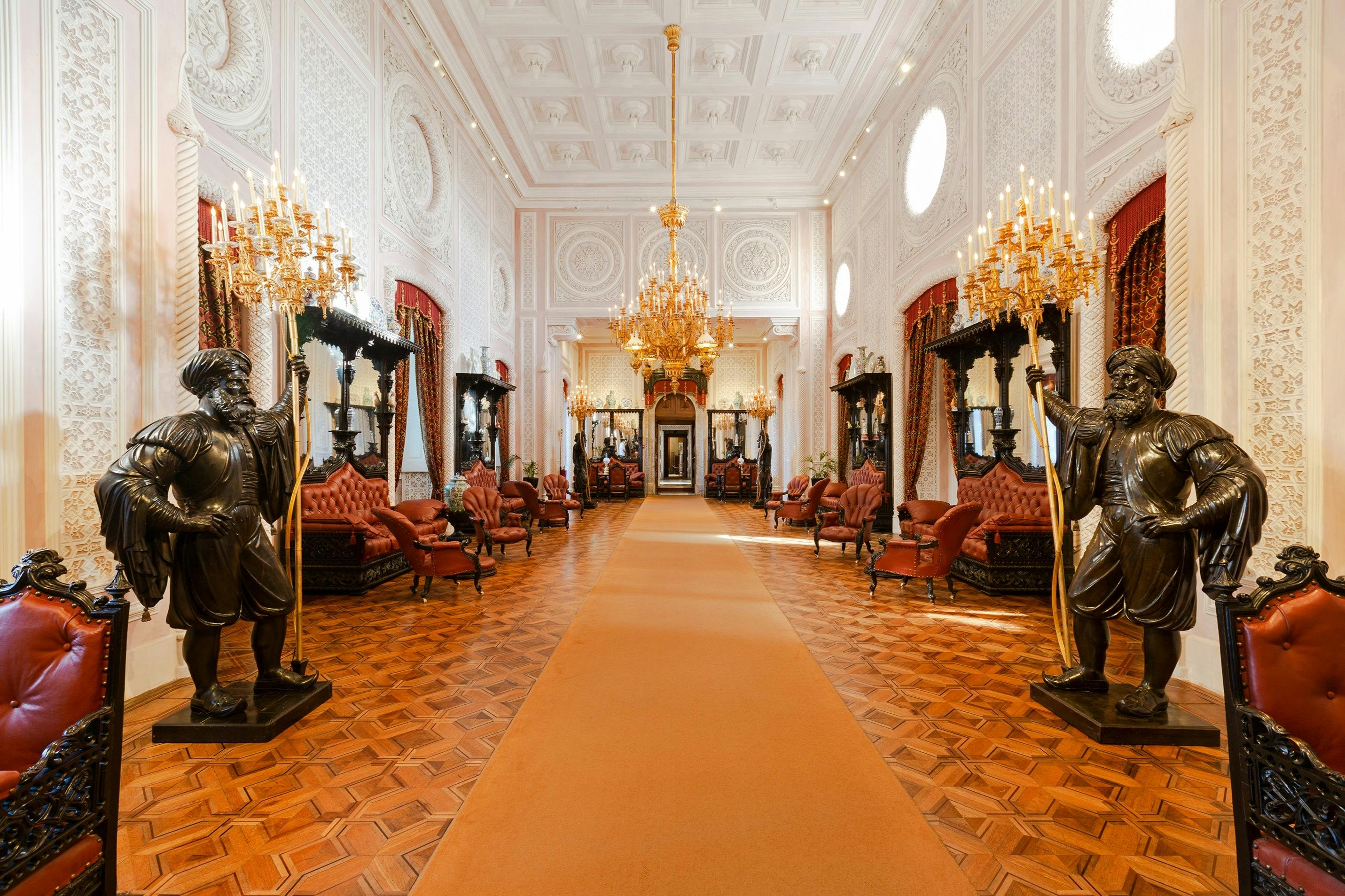 Sala ornata con lampadari, statue, divani rossi e un tappeto arancione su un pavimento in legno a motivi.