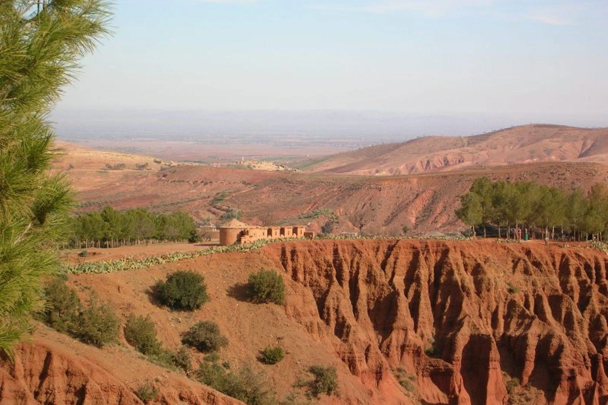 Arid landscape with red rocky terrain, sparse vegetation, and a cluster of buildings in the distance under a clear sky.