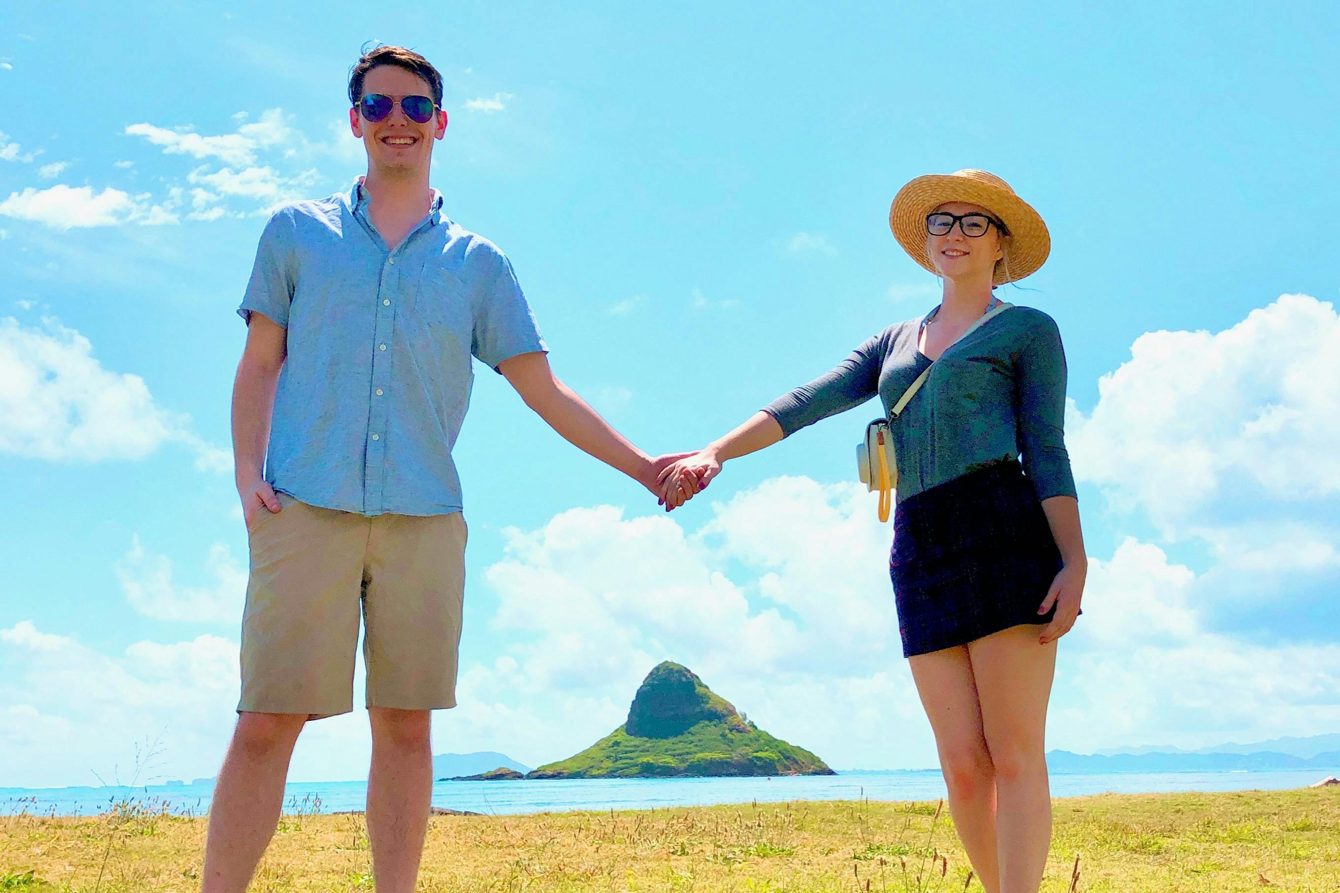 Two people holding hands in a sunny outdoor setting, with a grassy field, ocean, and a small island in the background.
