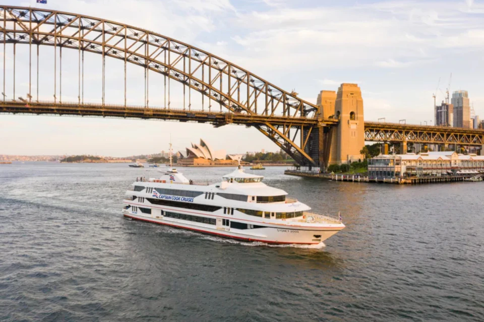 A large white cruise boat sails on a harbor with a bridge and the Sydney Opera House in the background.