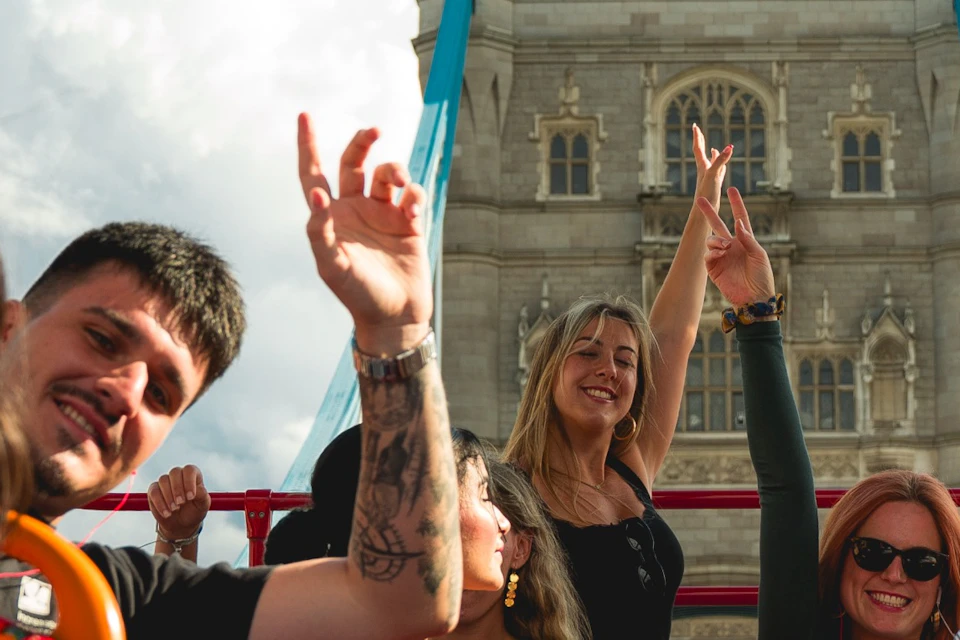 A group of people smiling and raising hands on a red open-top tour bus near Tower Bridge on a sunny day.