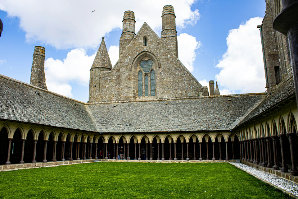 Courtyard of a historic stone building with arched walkways, tall chimneys, and green grass under a blue sky with white clouds.