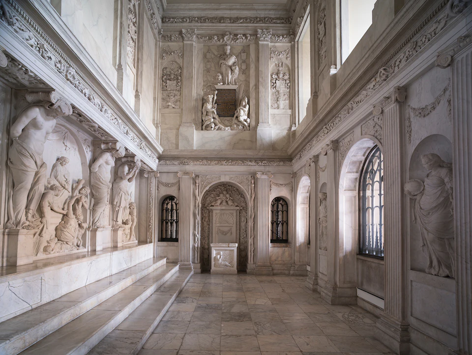 An ornate interior with marble steps, intricately carved walls, arched windows, and statues among detailed architectural elements.