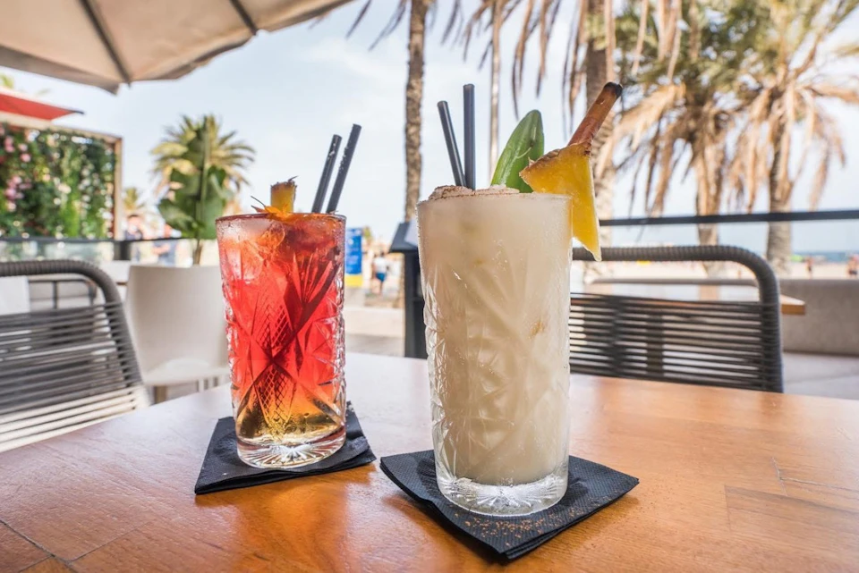 Two decorative cocktails on a table, one red and one white, garnished with fruit and spices, with a beach setting in the background.