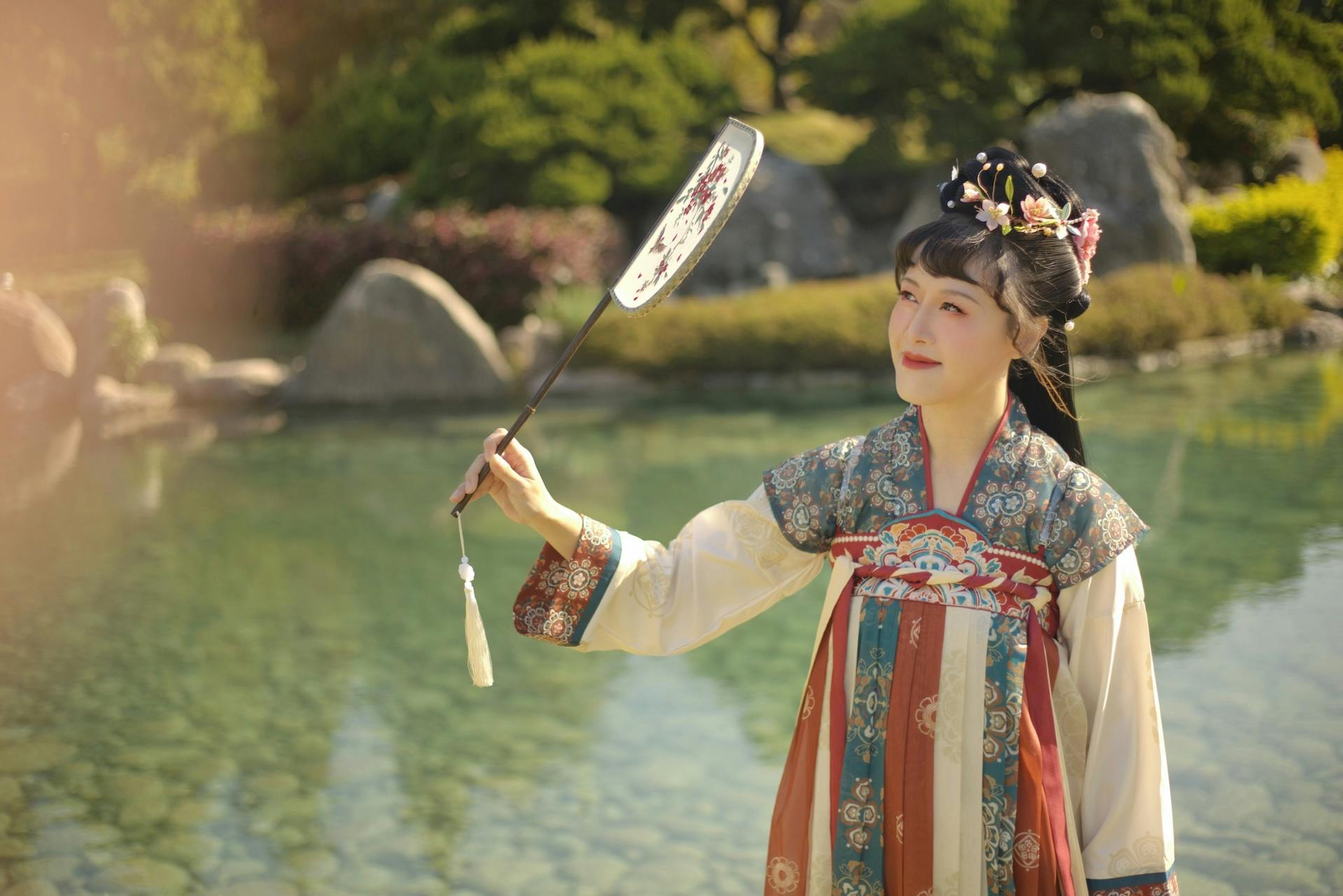 A person dressed in traditional clothing holding a decorated fan, standing near a pond in a lush garden.