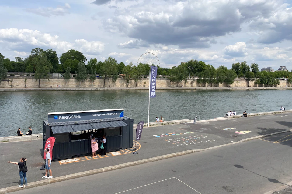 A riverside booth with "PARIS SEINE" branding, people nearby, and a river with trees in the background under a partly cloudy sky.