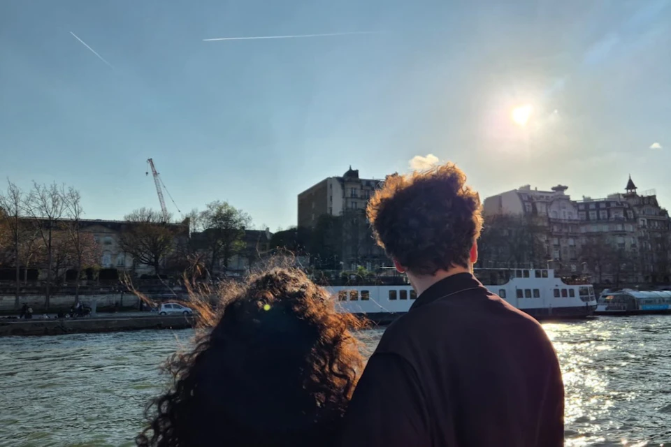 Two people with curly hair stand by a river under a sunny sky, with buildings, trees, and a crane in the background.