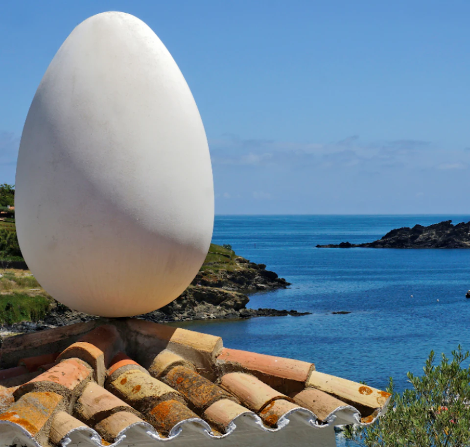 Large white egg sculpture on a tiled rooftop with a coastal landscape and sea in the background.