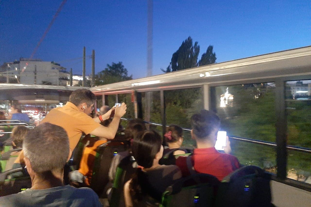 Ενήλικες και παιδιά κάθονται στο πάνω κατάστρωμα ενός τουριστικού λεωφορείου το σούρουπο, ενώ ένας άντρας με πορτοκαλί πουκάμισο βγάζει φωτογραφίες.