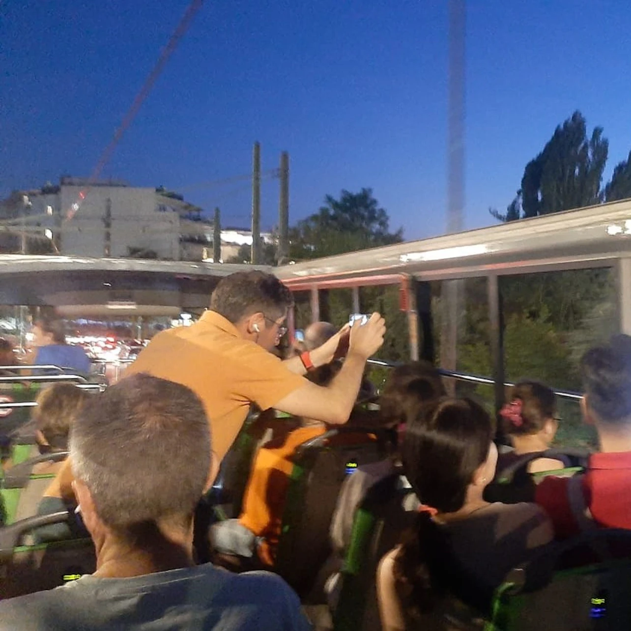 Adults and children sitting on the upper deck of a tour bus at dusk while a man in an orange shirt takes photos.