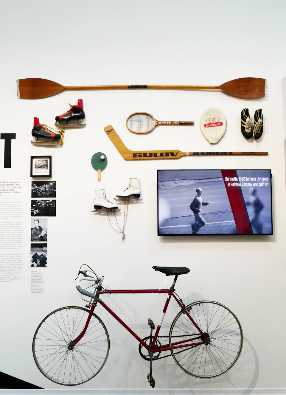A wall displaying sports equipment, including a wooden paddle, tennis racket, ice skates, and a bike, with a television screen showing a sports event.