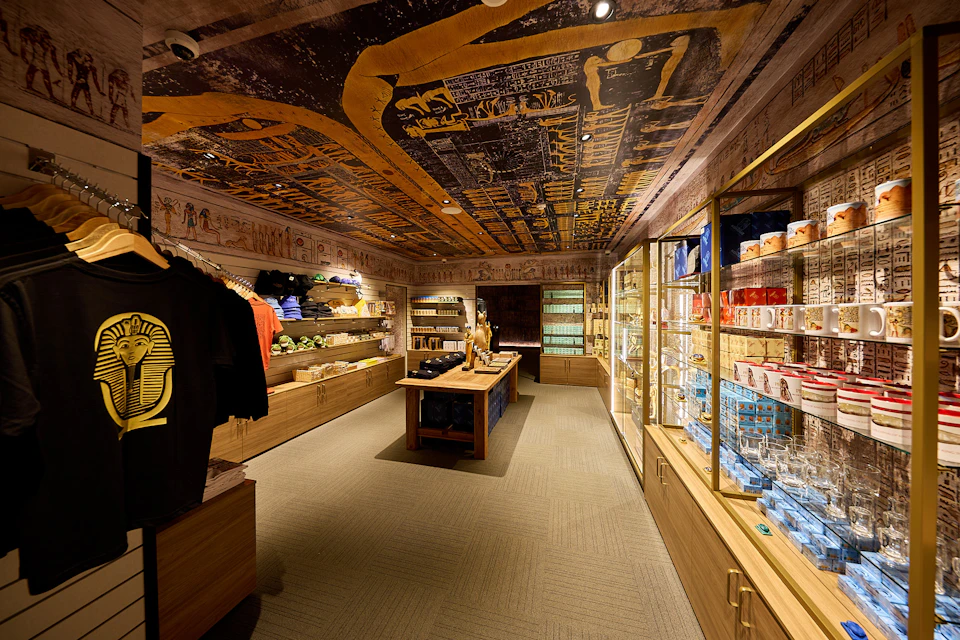 Gift shop with Egyptian-themed decorations, featuring shelves with clothing, souvenirs, and displays of various items under warm lighting.