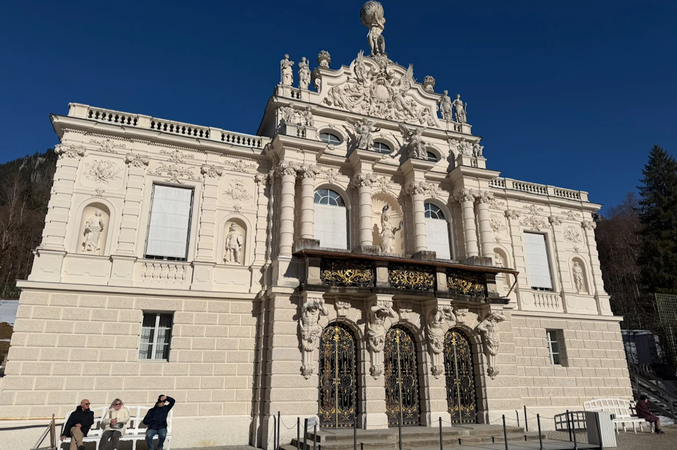 A baroque-style building facade with intricate sculptures, ornate doors, and a clear blue sky in the background.