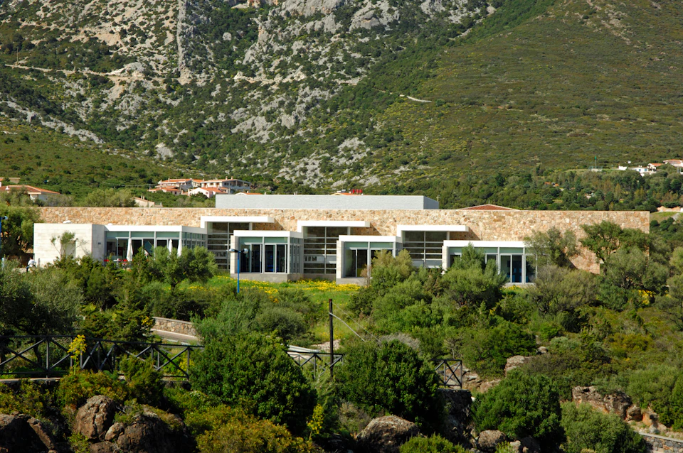A modern building with large windows sits at the base of a rocky, green hillside, surrounded by lush vegetation.