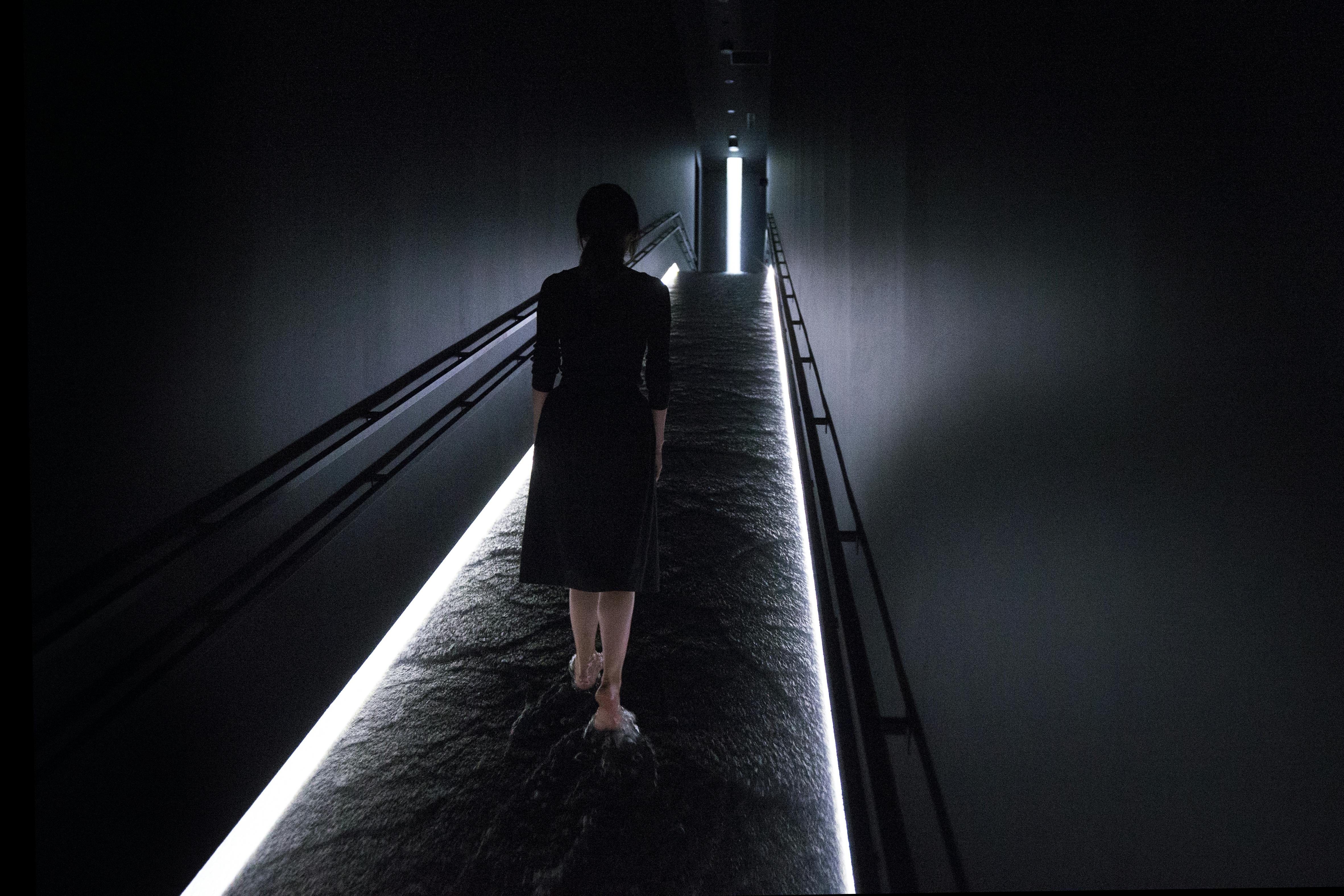 A person walks barefoot in a dimly lit hallway with water on the floor, illuminated by strip lighting along the edges.