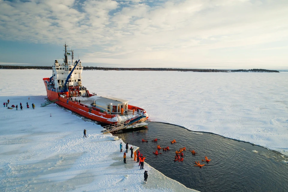 A red icebreaker ship docked on ice near an open water hole where people in orange suits are floating. Others stand on the ice.
