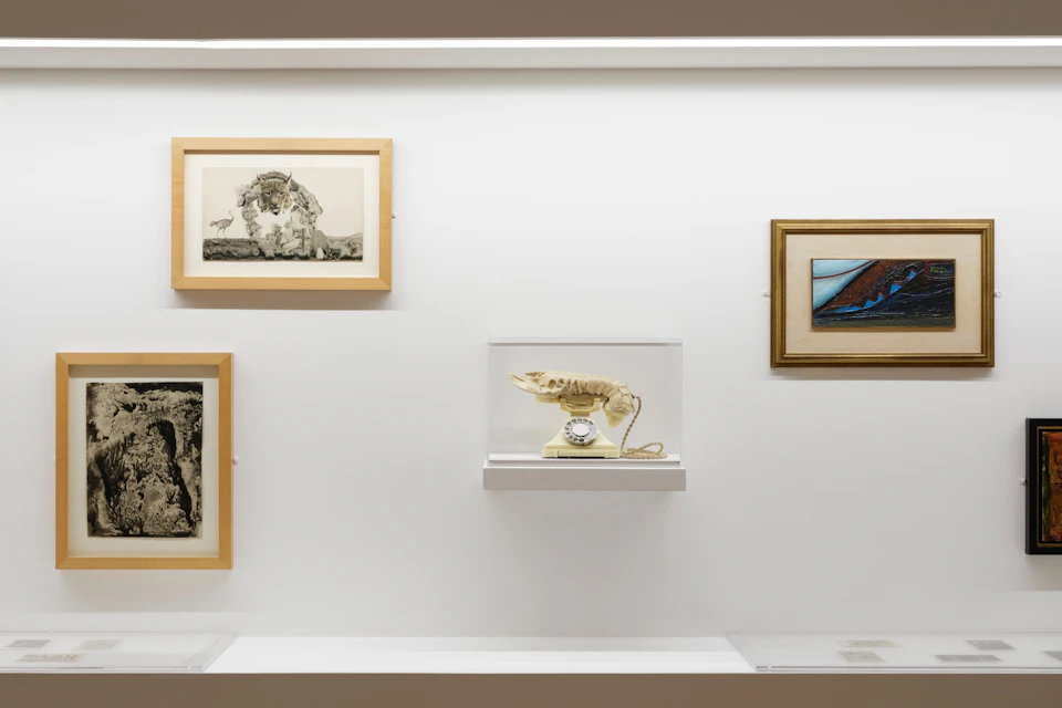 An art exhibit featuring framed artwork and a sculptural lobster telephone displayed on a white wall.