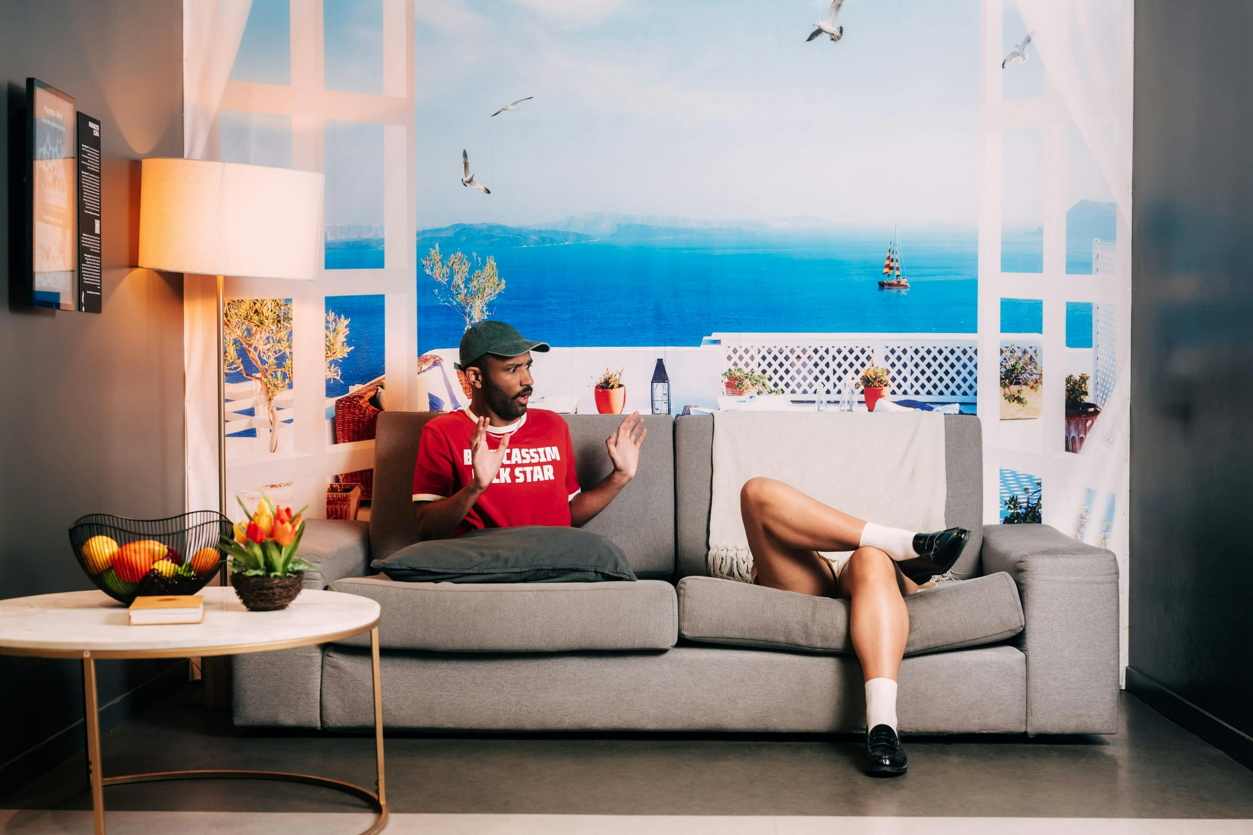 海岸をテーマにした背景、近くに花やフルーツが置かれたサイドテーブルがある部屋で、灰色のソファに座っている2人。