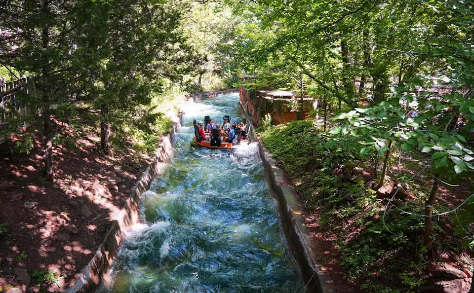 People riding a circular raft on a winding water rapids ride surrounded by lush green trees and foliage.
